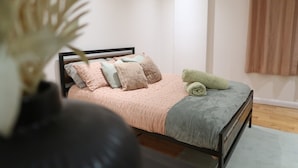 Una cama cuidadosamente tendida con una colcha rosa, almohadas, un plaid gris y una toalla verde sobre un cabecero de madera.