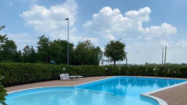 En swimmingpool med flisekant, omgivet af en lav hæk og en belagt terrasse med liggestole.