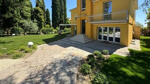 A two-story yellow house with a balcony, a paved driveway, and a well-maintained garden with trees and shrubs. - La Gialla (Attico d'Amore) By Case al Lago (Padenghe sul Garda)