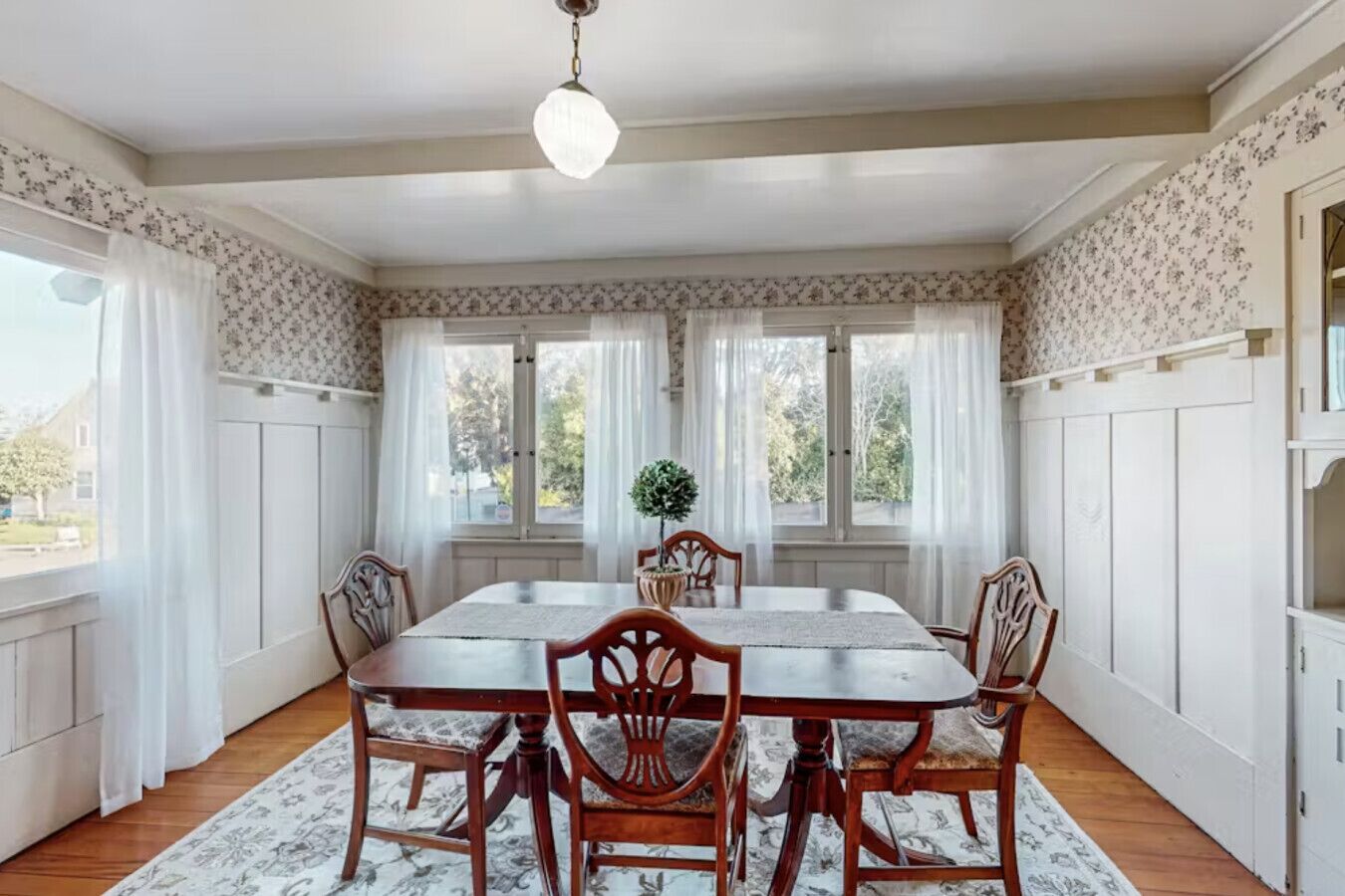 Une salle à manger avec une grande table, six chaises, un lustre et une vue sur l’extérieur grâce à de grandes fenêtres.