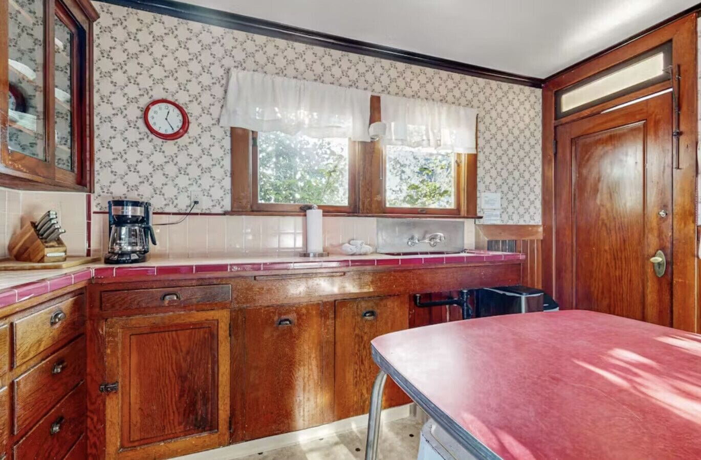 Een keuken met houten kastjes, een rode aanrecht, een spoelbak, een raam met gordijnen en een klok aan de muur.