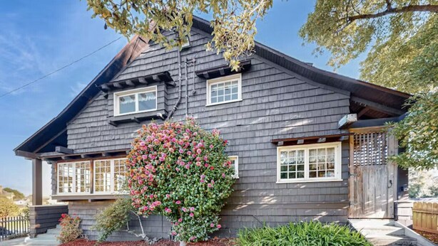 A two-story house with a dark shingled exterior, a front porch, and a large flowering bush.