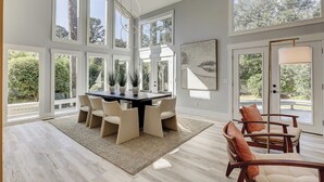 A spacious dining area with a large table and multiple chairs, surrounded by large windows offering views of greenery. - Sunset Rentals | Red Oak 40 (Hilton Head Island)