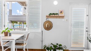 Een kamer met een houten tafel en stoelen, een witte muur met een plank waarop planten en een hoed staan, en uitzicht op zee door open luiken.
