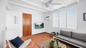 Een woonkamer met een witte plafondventilator, een houten deur, een tv op een wit kastje, een bank en een bijzettafeltje met een plant.