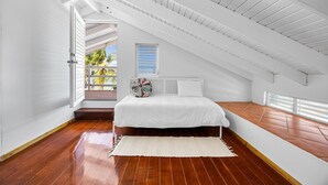 Een slaapkamer met een schuine plafond, een bed met witte beddengoed, een kleurrijk kussen, een raam met jaloezieën en uitzicht op palmbomen.