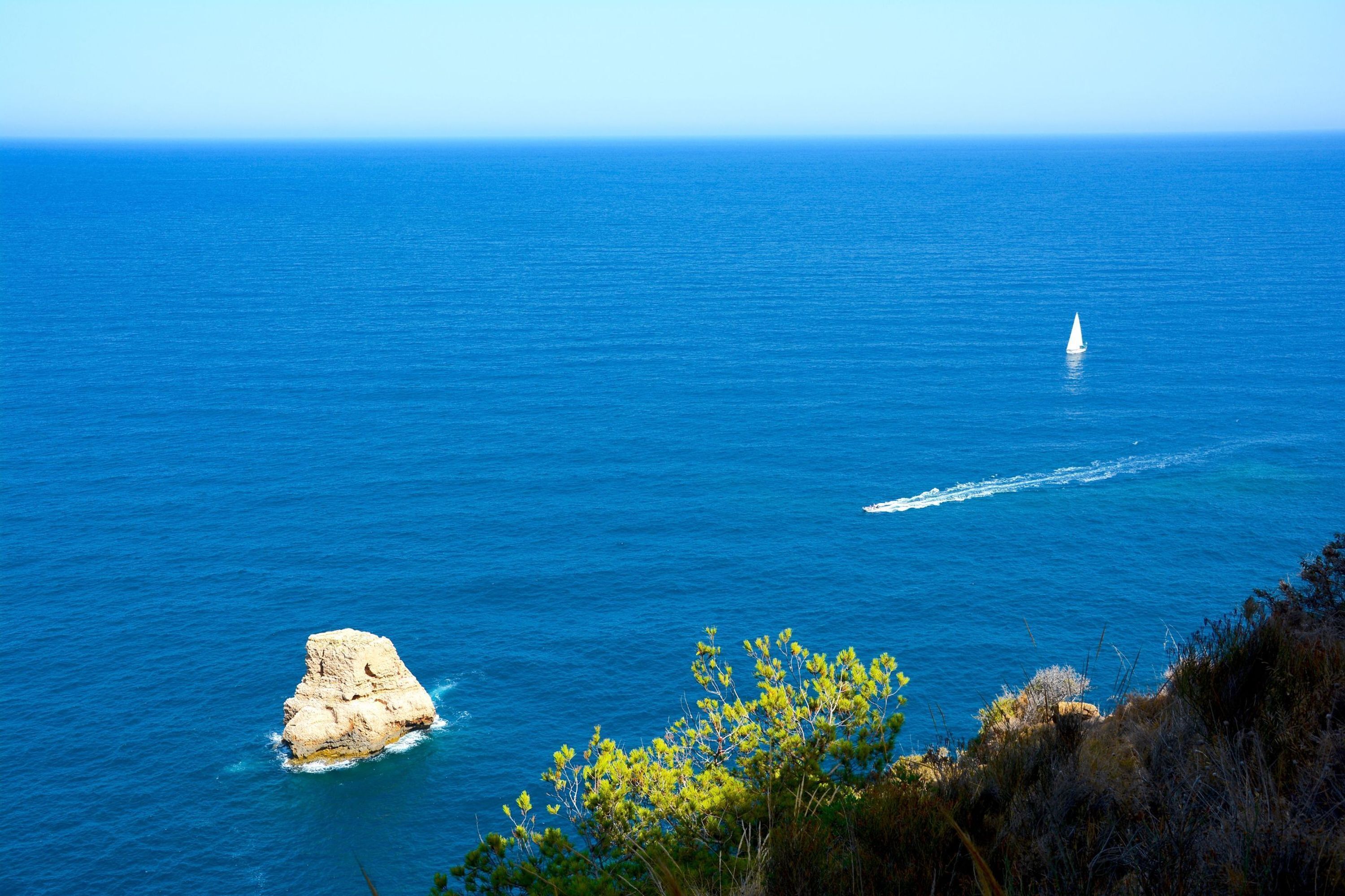 Paisaje costero con vista despejada al mar, una pequeña formación rocosa y un velero a lo lejos.