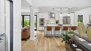 A modern kitchen with white cabinetry, a marble backsplash, and stainless steel appliances. There are bar stools at the island and a dining area with a large window. - Sunset Rentals | Planters Wood 48 (Hilton Head Island)