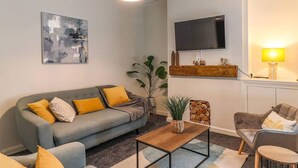 A living room with a grey sofa, yellow cushions, a wooden coffee table, a TV mounted on the wall, and a small plant on the floor. - Cardiff Gem on Topaz Street - 4 Bedroom House (Cardiff)