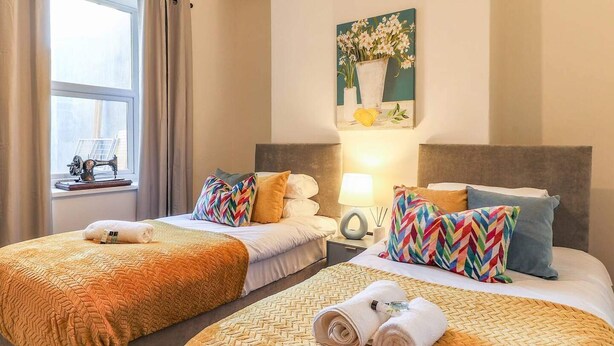 A hotel room with two beds, a painting above the beds, a lamp on the nightstand, and a sewing machine on a table by the window. - Cardiff Gem on Topaz Street - 4 Bedroom House (Cardiff)