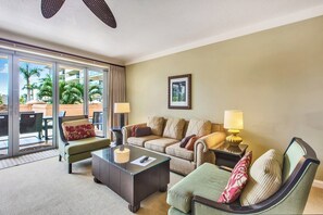 Ein Hotelzimmer mit Sofa, Sessel und einem Glastisch. Ein Schiebetür öffnet sich zu einem Außenbereich mit Blick auf Palmen.
