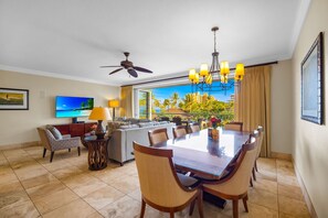 A hotel room with a dining area, a sofa, a TV, and a view of palm trees and buildings. - Honua Kai | Ocean View 10 BR, Sleeps 28 | Car Incl. w/ 6+ Nights | HKH ML-1105 by KBM (Lahaina)