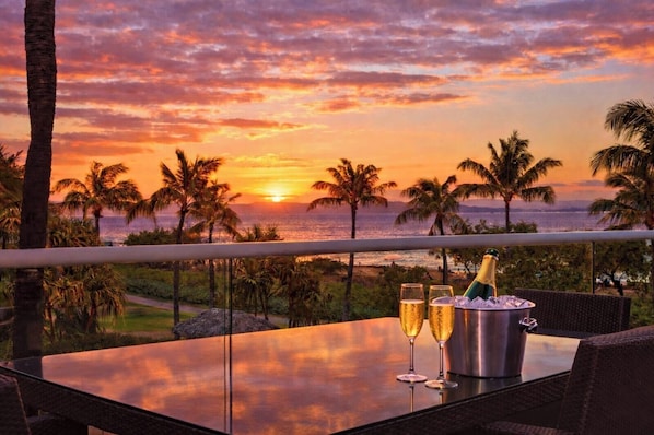 Une table avec deux coupes de champagne et une bouteille, donnant sur un coucher de soleil avec des palmiers et l’océan.