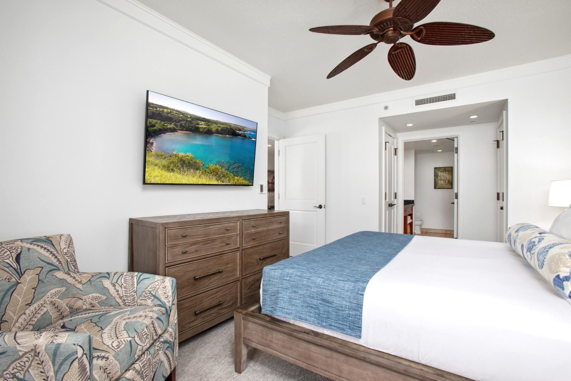 Een slaapkamer met een bed, een kaptafel, een stoel, een plafondventilator en een landschapsschilderij.