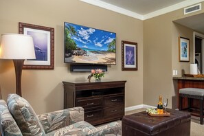 Ein Wohnzimmer mit Flachbildfernseher, einer dunklen Holz-Unterhaltungseinrichtung, einem gemusterten Sofa, einem braunen Lederhocker und einem gerahmten Bild von einer Strandlandschaft.