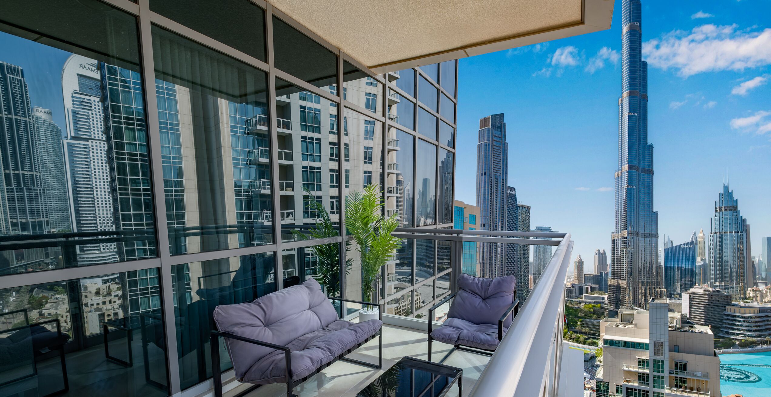 A modern balcony with a sofa and a small table, overlooking a cityscape with tall buildings and a clear sky.