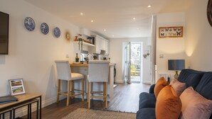 A modern living room with a dark blue sofa, two light grey chairs, and a patterned rug. A kitchen with white cabinets and a dining area with a small table and chairs is visible in the background. - Eldred Cottage, Middleham (Middleham, North Yorkshire)