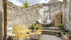 A stone-walled patio with yellow wicker furniture, a small table with a plant, and a wooden door with a heart decoration. - Eldred Cottage, Middleham (Middleham, North Yorkshire)