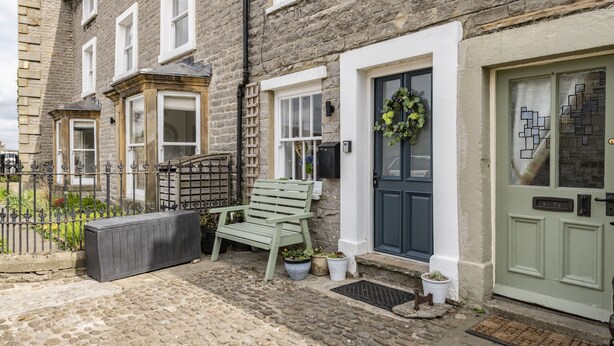 A stone building with a green bench, potted plants, and a door with a wreath. - Eldred Cottage, Middleham (Middleham, North Yorkshire)