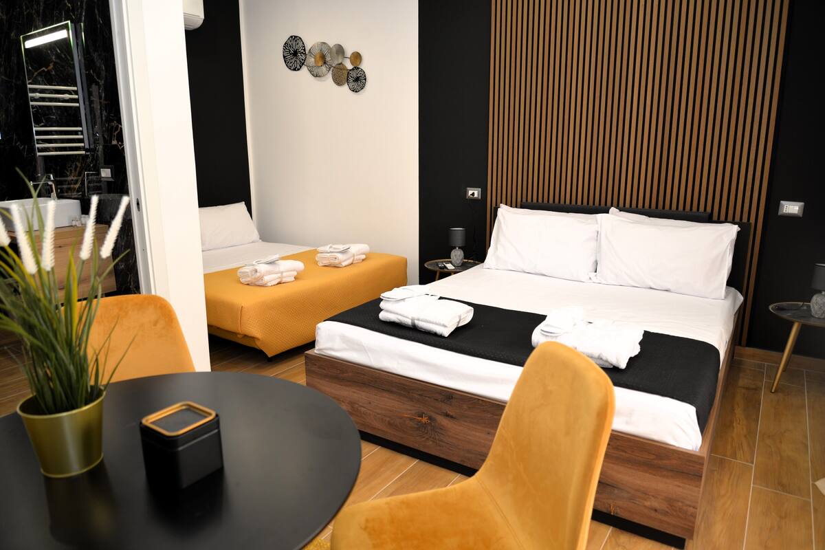 A modern hotel room with a large bed, a smaller sofa, a round table, and a potted plant.
