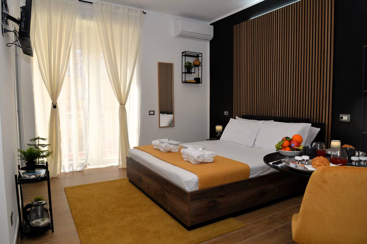 A modern hotel room with a wooden bed, a bedside table with a lamp, a small plant, and a tray with breakfast items.