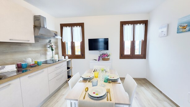 Une cuisine compacte avec des meubles blancs, une cuisinière intégrée et un coin repas aménagé pour quatre personnes, comprenant une table, des chaises et un téléviseur fixé au mur.