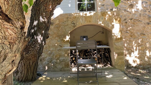 Un área de estar rústica al aire libre bajo un gran árbol, con un muro de piedra y una ventana al fondo.