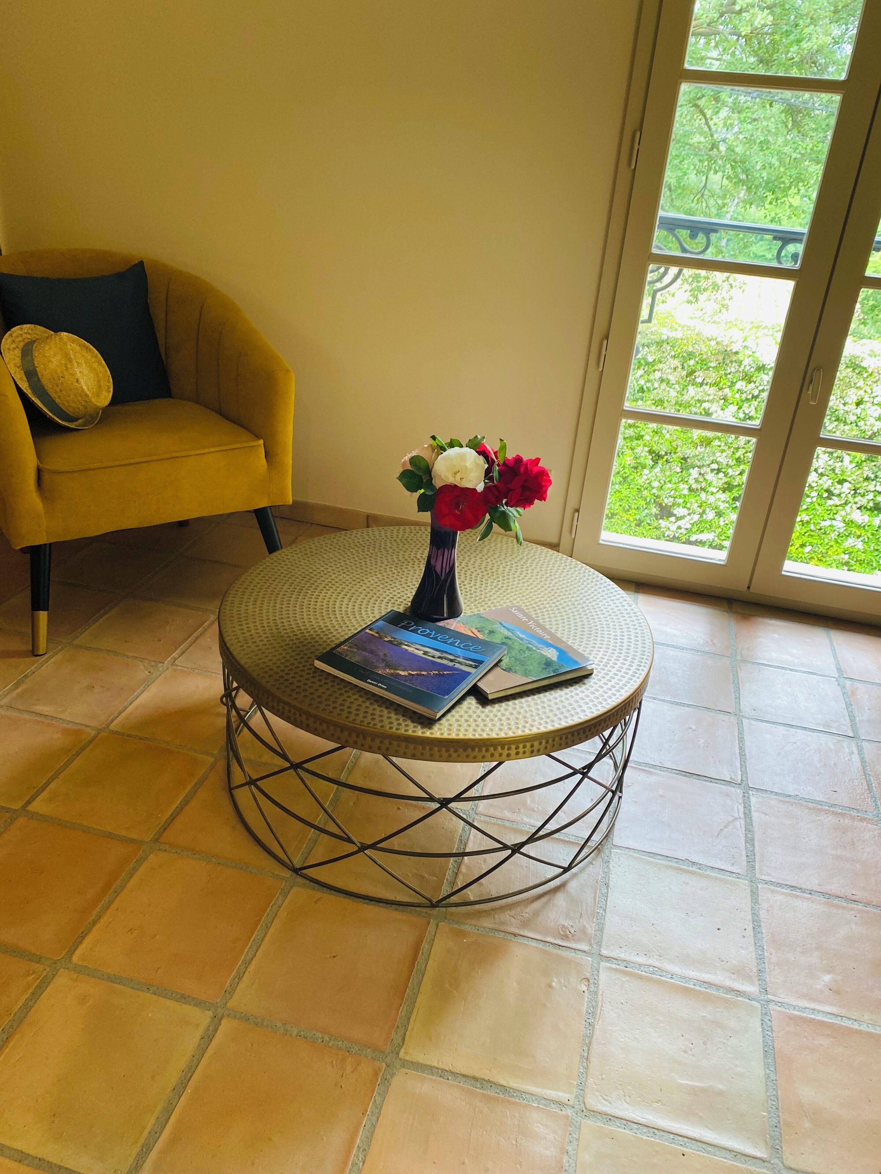 Un fauteuil jaune surmonté d’un chapeau, une table ronde avec un livre, et un vase rempli de fleurs.