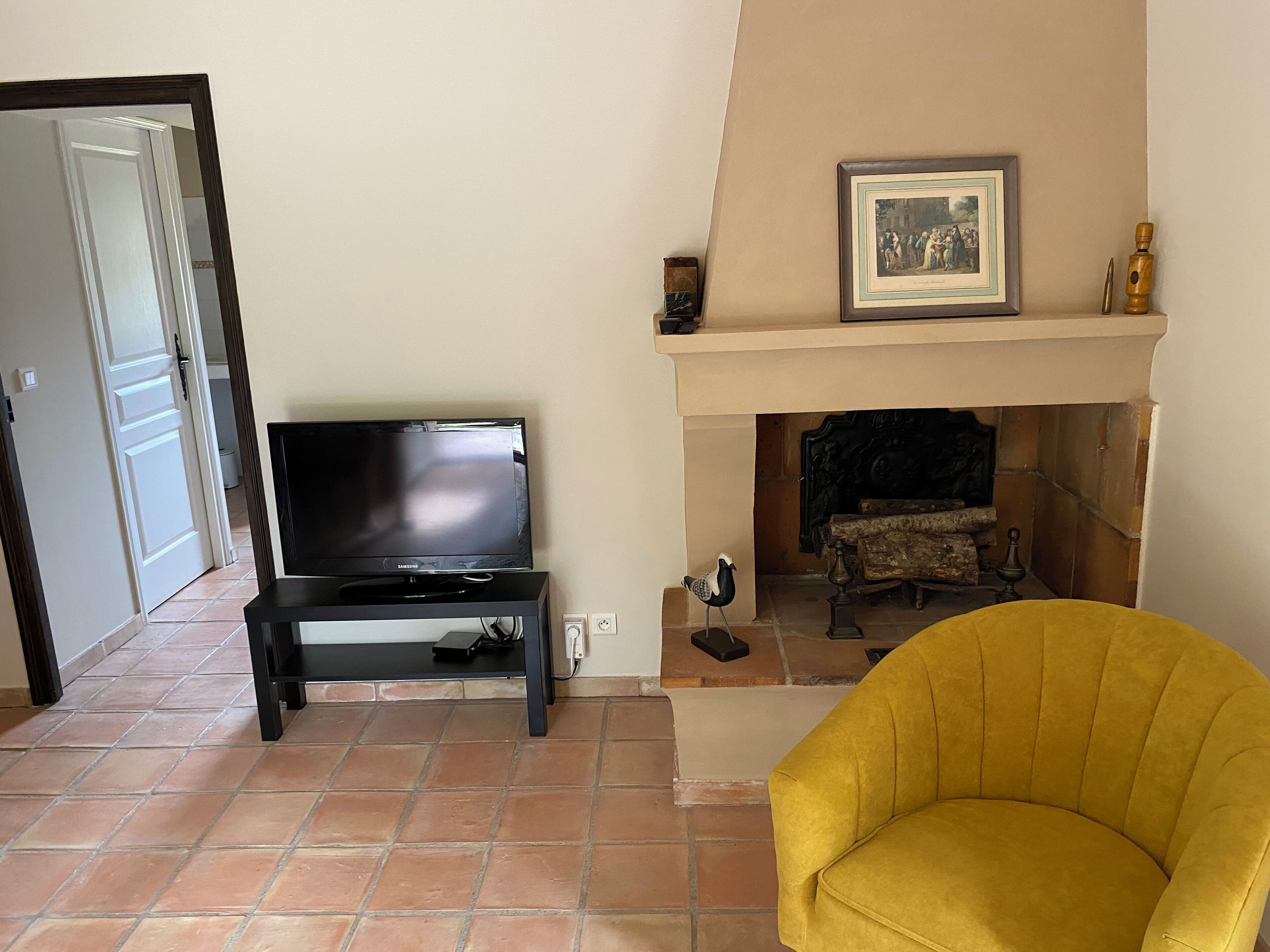 A living room with a television on a stand, a yellow armchair, a fireplace, and a framed picture above the mantel.