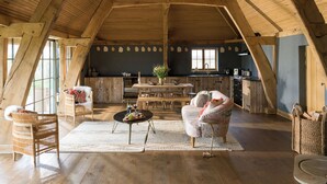 A spacious loft with a high ceiling, exposed wooden beams, a dining area, and a kitchen with a large window.