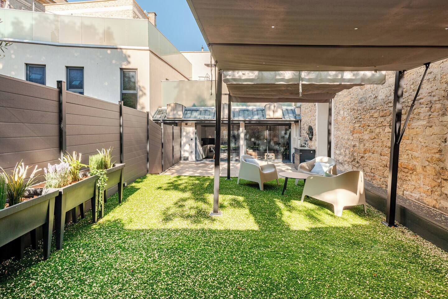 Un espace extérieur moderne, aménagé en patio, avec un coin salon abrité, un petit jardin planté et un mur en pierre.