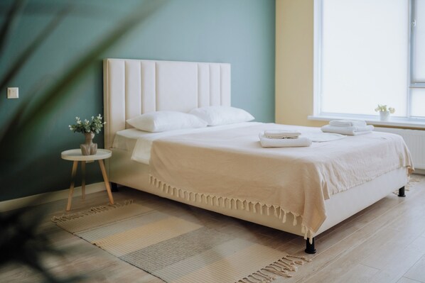 A neatly made bed with a white headboard and pillows, a small bedside table with a vase of flowers, and a window with a view of the outside.