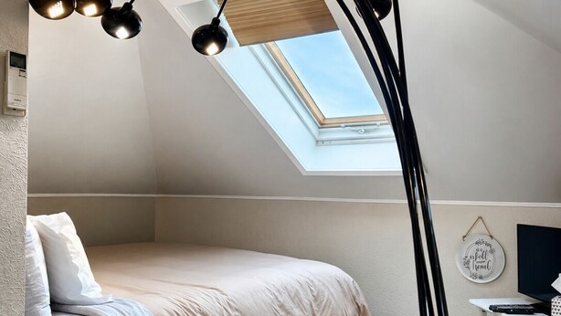 A bedroom with a bed, a desk, a black floor lamp, a skylight, and a wall-mounted thermostat.