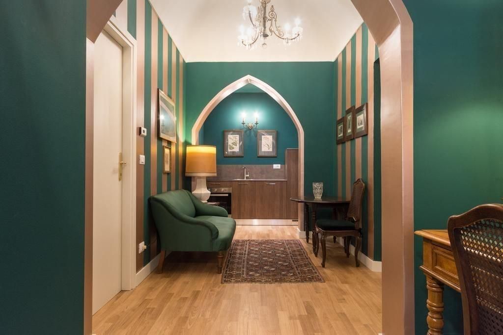 A hallway with a green wall, wooden flooring, a green armchair, a small table with chairs, and a kitchen area with wooden cabinets and a chandelier.
