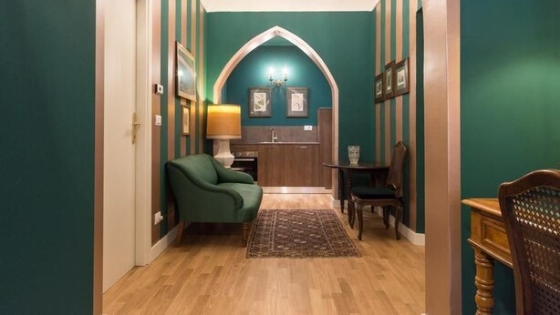 A hallway with a green wall, wooden flooring, a green armchair, a small table with chairs, and a kitchen area with wooden cabinets and a chandelier. - Porta Palace Apartments (Turin)