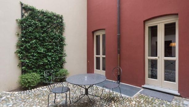 A small outdoor patio with a round table and two chairs, a wall covered with climbing plants, and a red building with a white door and window. - Porta Palace Apartments (Turin)