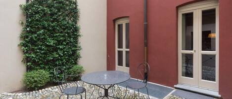 Une petite terrasse extérieure avec une table ronde et deux chaises, un mur recouvert de plantes grimpantes et un bâtiment rouge avec une porte et une fenêtre blanches.