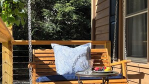 A wooden swing bench with a cushion and a small table with a glass mug and fruit.