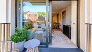 En modern hotellentrél med en glasdörr, en liten utesittgrupp med möbler i rotting och en utsikt över gatan och byggnaderna i bakgrunden.
