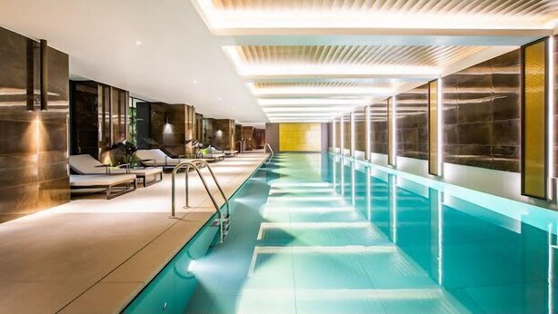A long indoor swimming pool with a modern design, including a ceiling with linear light fixtures and a poolside area with lounge chairs. - Contemporary Marylebone with Pool and Gym AC (Greater London)