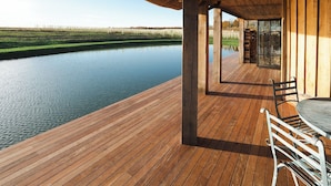 Une terrasse en bois surmontée d’un toit de chaume, offrant une vue sur un plan d’eau paisible et un champ verdoyant.
