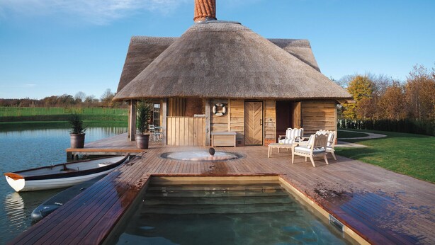 Un bâtiment à toit de chaume doté d’un jacuzzi et de chaises longues, offrant une vue sur un lac paisible.