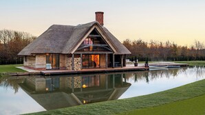 Une maison au toit de chaume, dotée d’une grande porte vitrée, d’une cheminée et dont le reflet se mire dans l’eau.