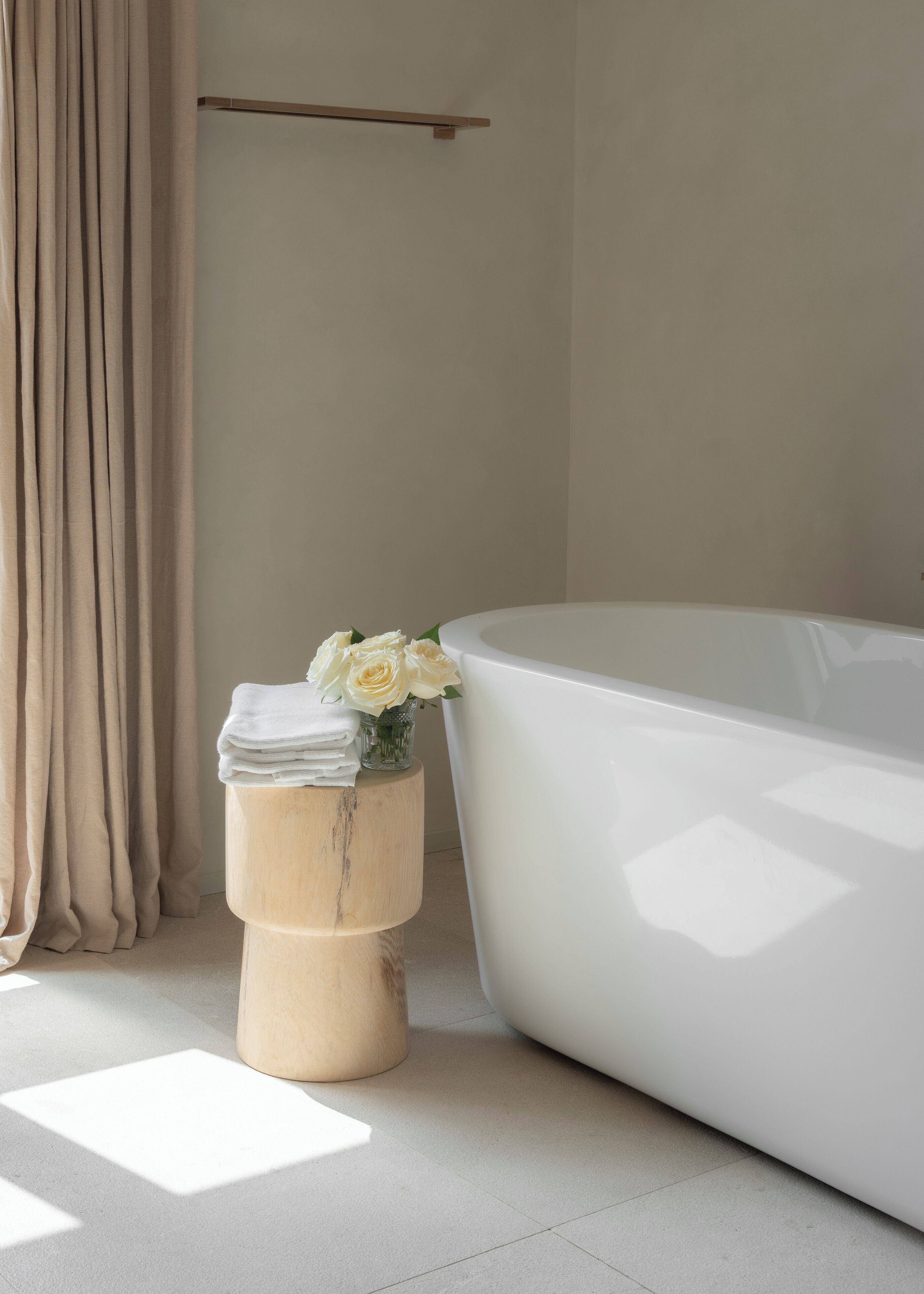 A modern bathroom with a white bathtub, a wooden stool with towels and a vase of white roses, and a beige curtain.