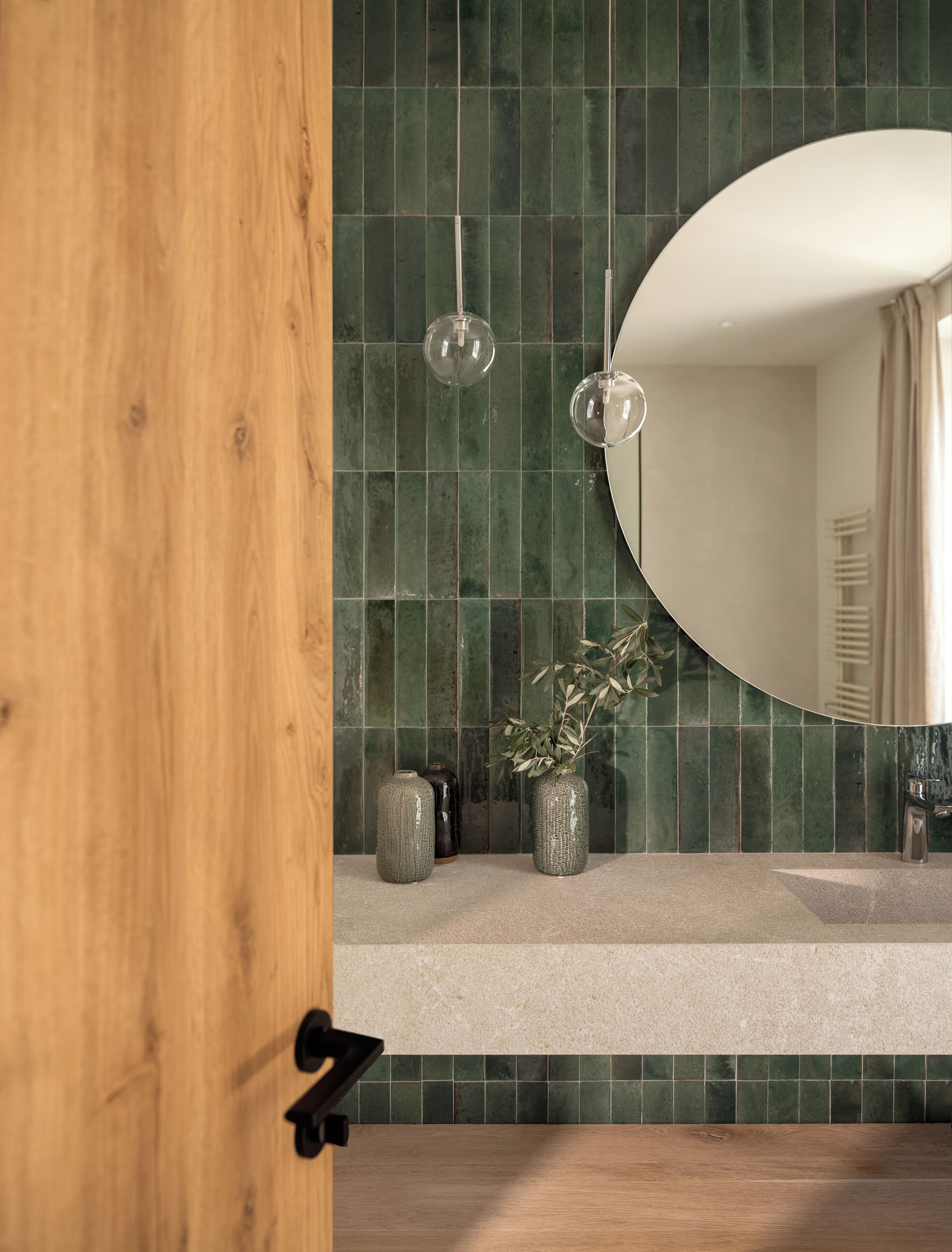 A bathroom with a wooden door, green tiled walls, a round mirror, and two hanging light fixtures.