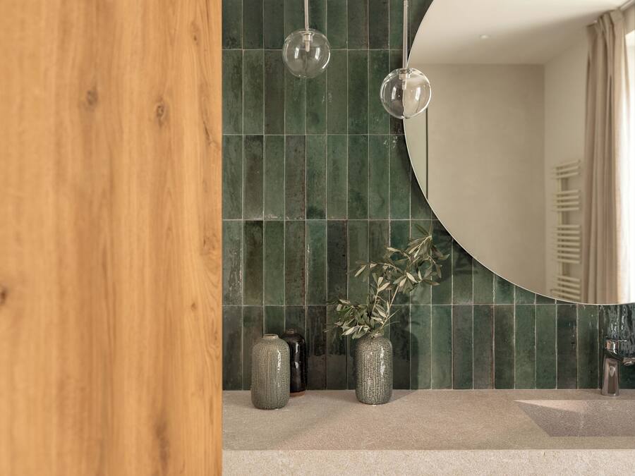 A bathroom with a wooden door, green tiled walls, a round mirror, and two hanging light fixtures.