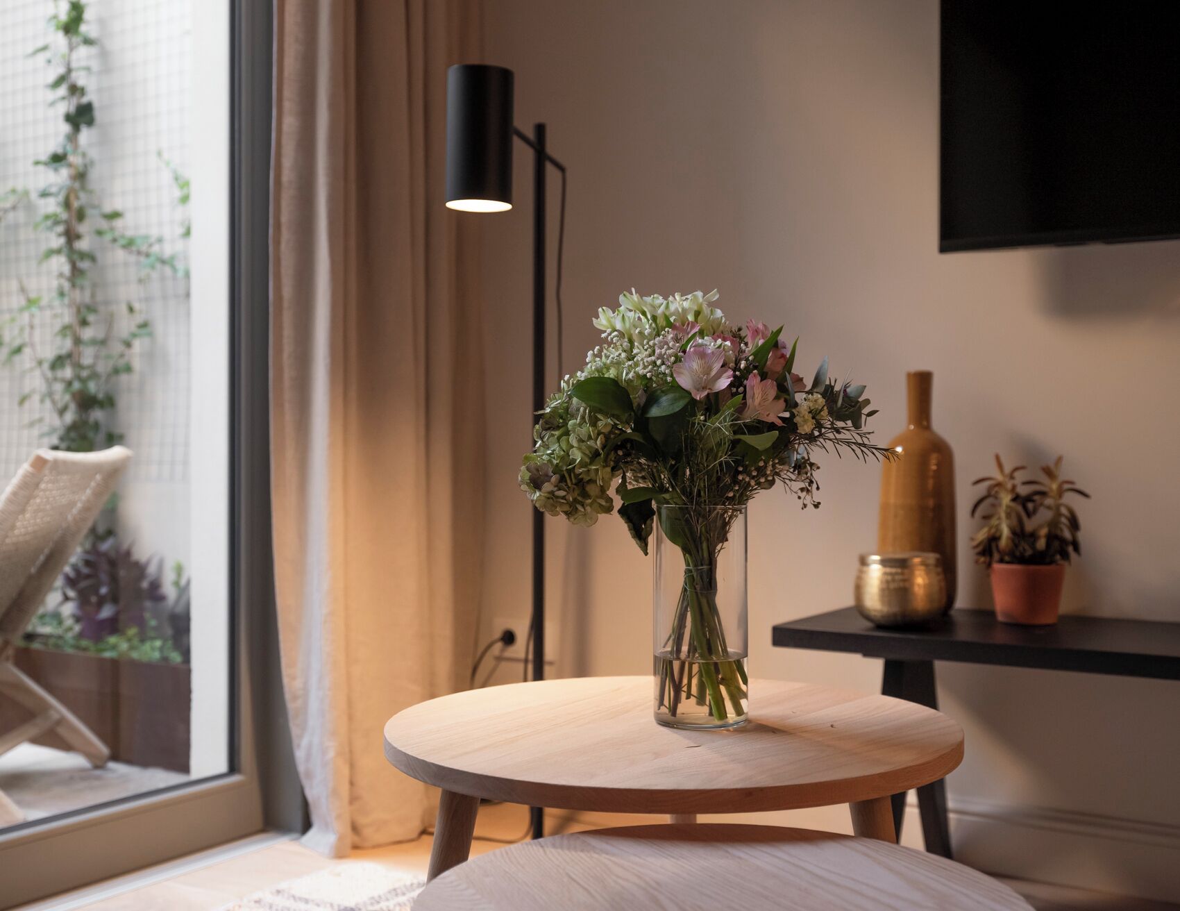 A round wooden table with a clear glass vase containing flowers, a black floor lamp, and a television mounted on the wall.