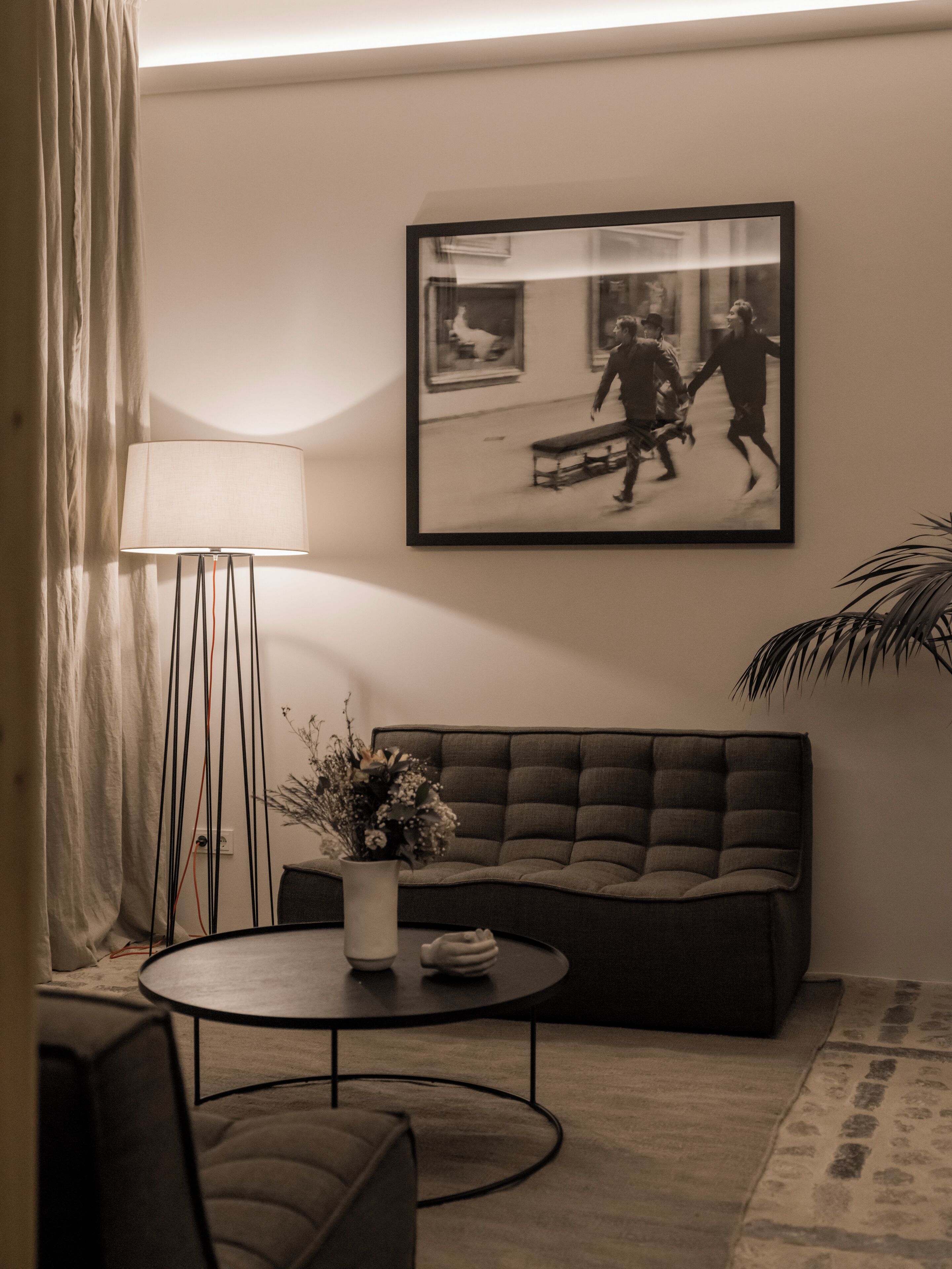 Een woonkamer met een bank, een rond bijzettafeltje, een vloerlamp en een zwart-wit foto in een lijst aan de muur.