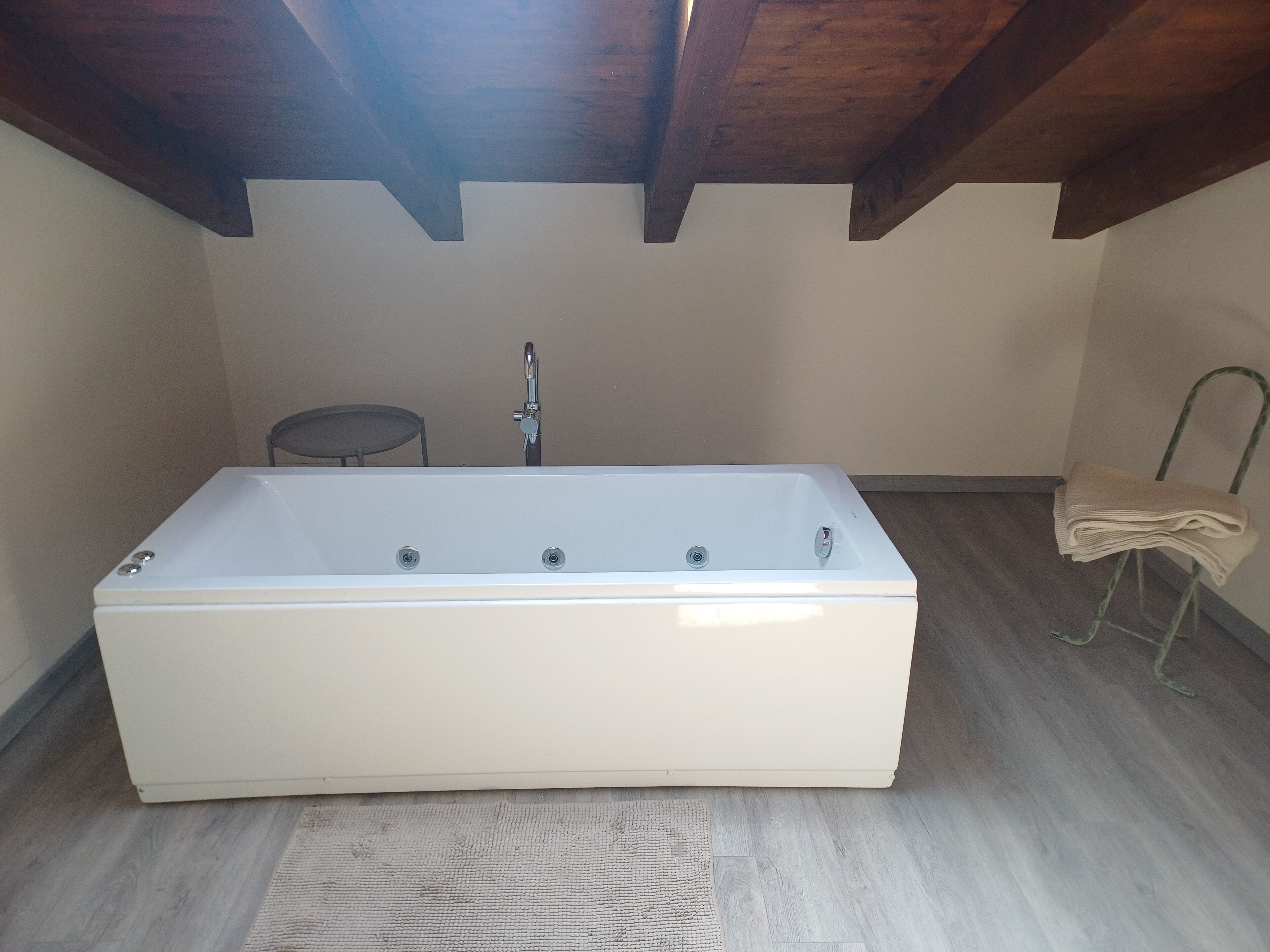 Une baignoire rectangulaire dans une pièce dont le plafond est orné de poutres en bois, une petite table ronde et une chaise sur laquelle est posée une serviette.