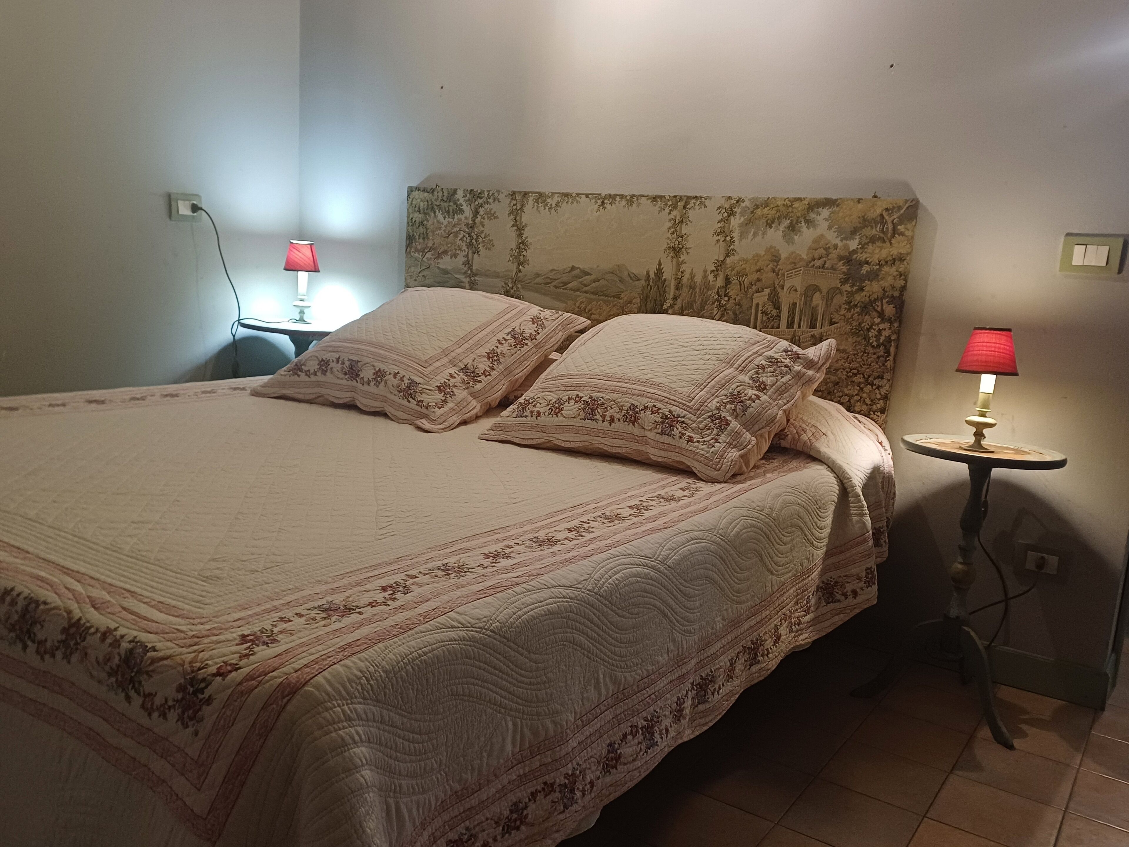 A neatly made bed with a floral-patterned bedspread and pillows, two bedside lamps, and a wall-mounted light switch.